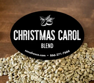 Close-up of Sonofresco Christmas Carol Blend label displayed above unroasted green coffee beans on a wooden surface.