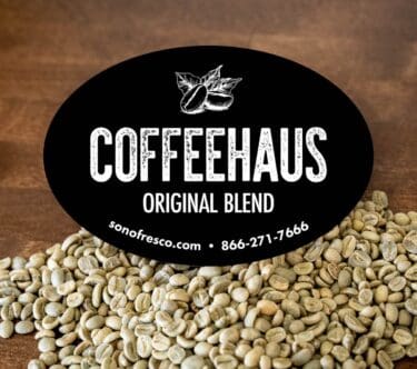 Black oval label reading ‘COFFEEHAUS Original Blend’ with a coffee bean illustration. Background shows green coffee beans on a wooden surface.